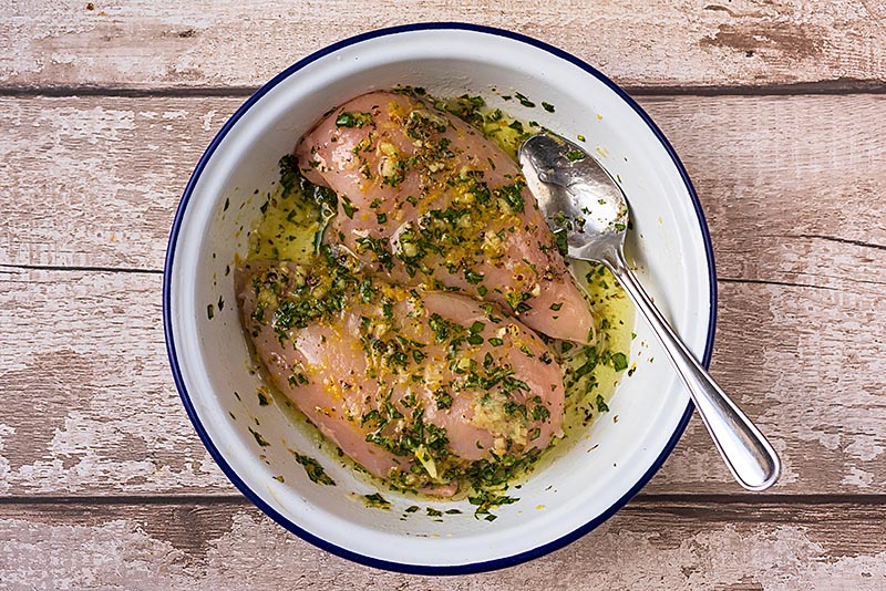 Two raw chicken breasts in a round dish with, lemon juice, zest, oil and chopped basil all mixed together.