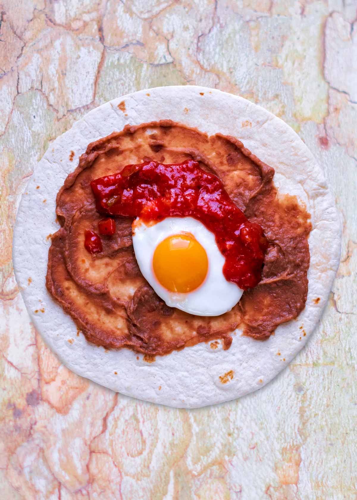 A flour tortilla spread with refried beans and a fried egg and salsa on top.