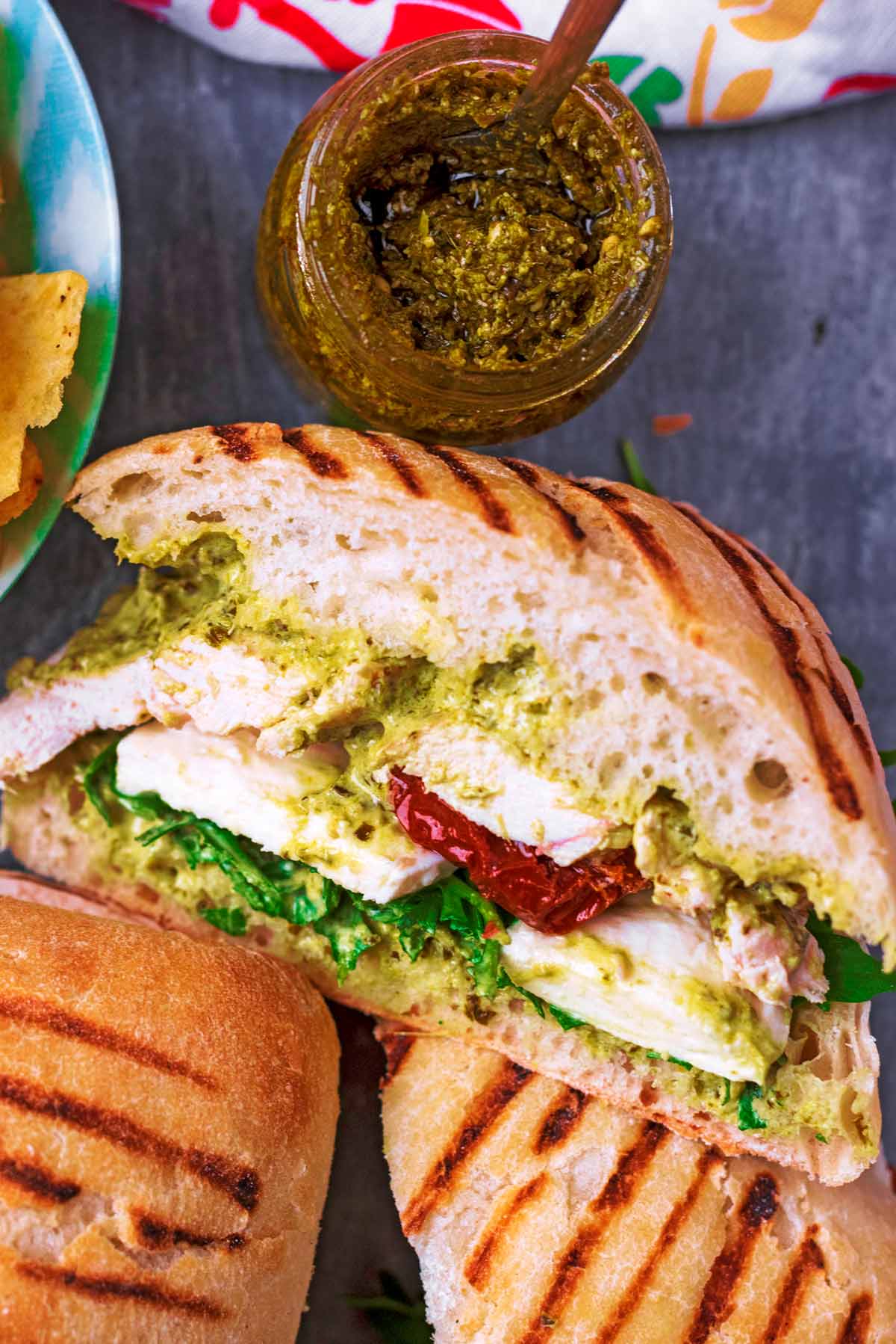 A ciabatta bun with chicken, lettuce, tomatoes and pesto sliced in half next to a jar of pesto.