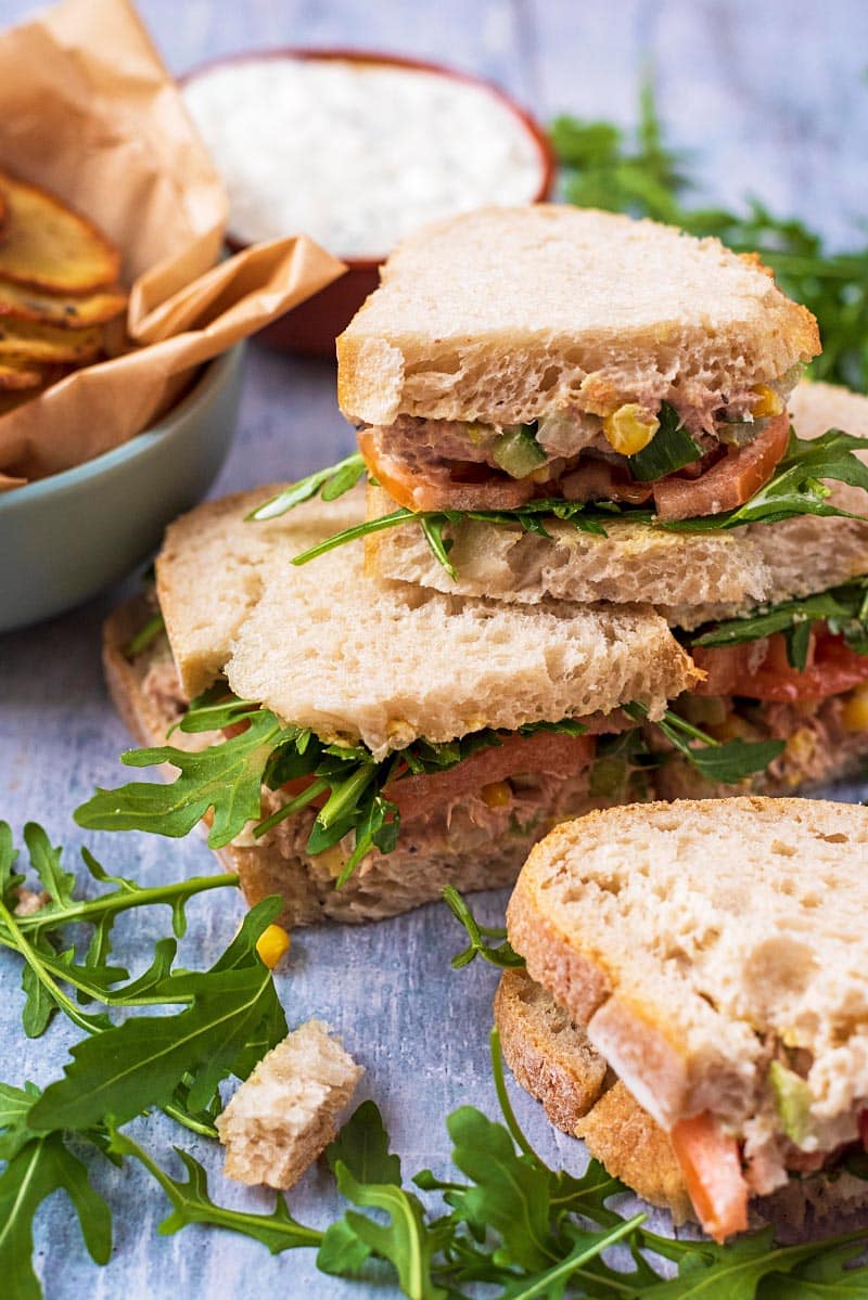 Four tuna sandwiches stacked up, surrounded by arugula and potato chips.