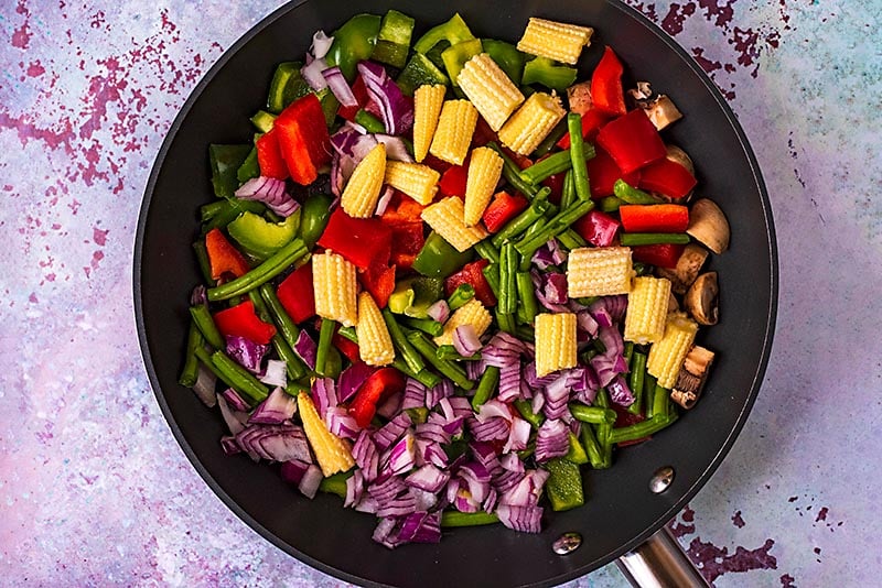 A frying pan containing chopped red onion, baby corn, green beans, bell peppers and mushrooms.