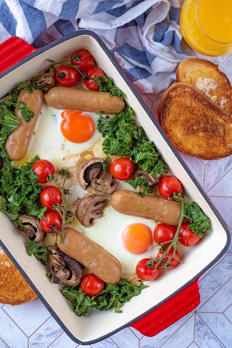 A large red dish containing eggs, tomatoes, mushrooms, kale and sausages.
