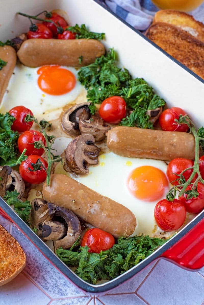 A large rectangular baking tray with sausages, eggs, tomatoes, kale and mushrooms.