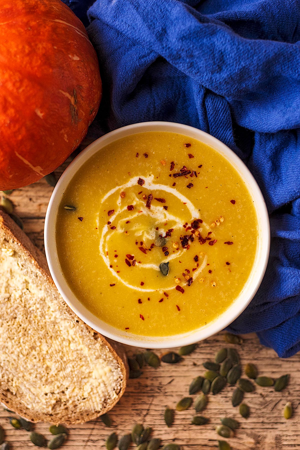 A bowl of pumpkin soup next to a whole pumpkin and a slice of buttered bread.