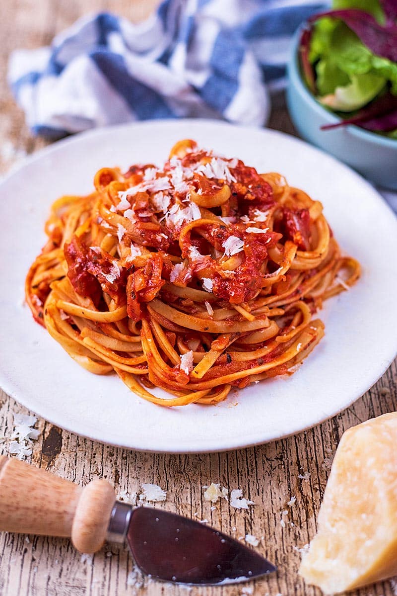 A plate of pasta in a tomato sauce next to a bowl of salad and a block of parmesan.