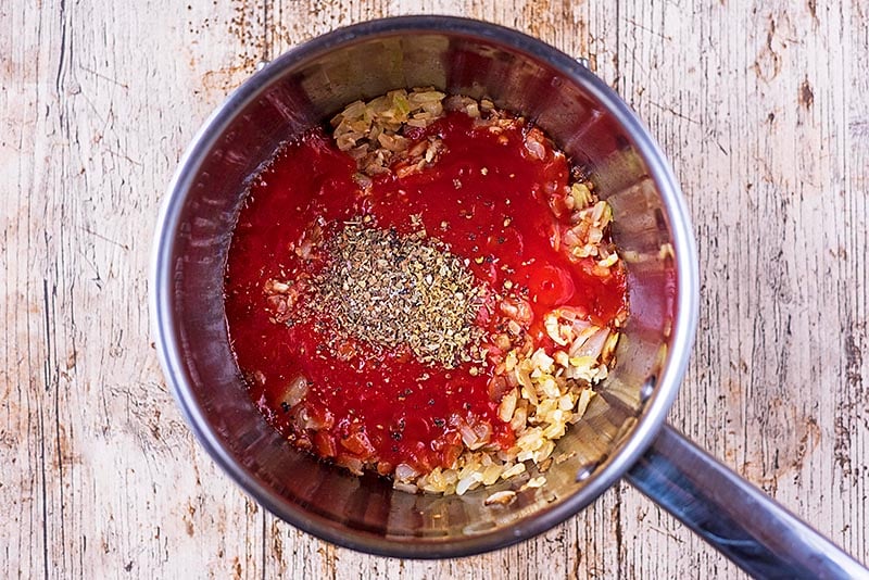 A saucepan containing chopped onions, minced garlic, passata and herbs.