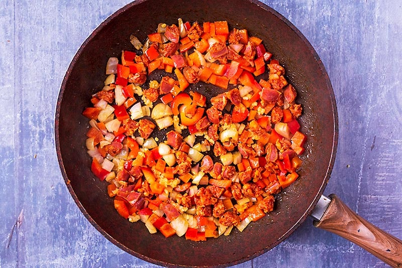 Chopped chorizo, red bell pepper and onion cooking in a frying pan.