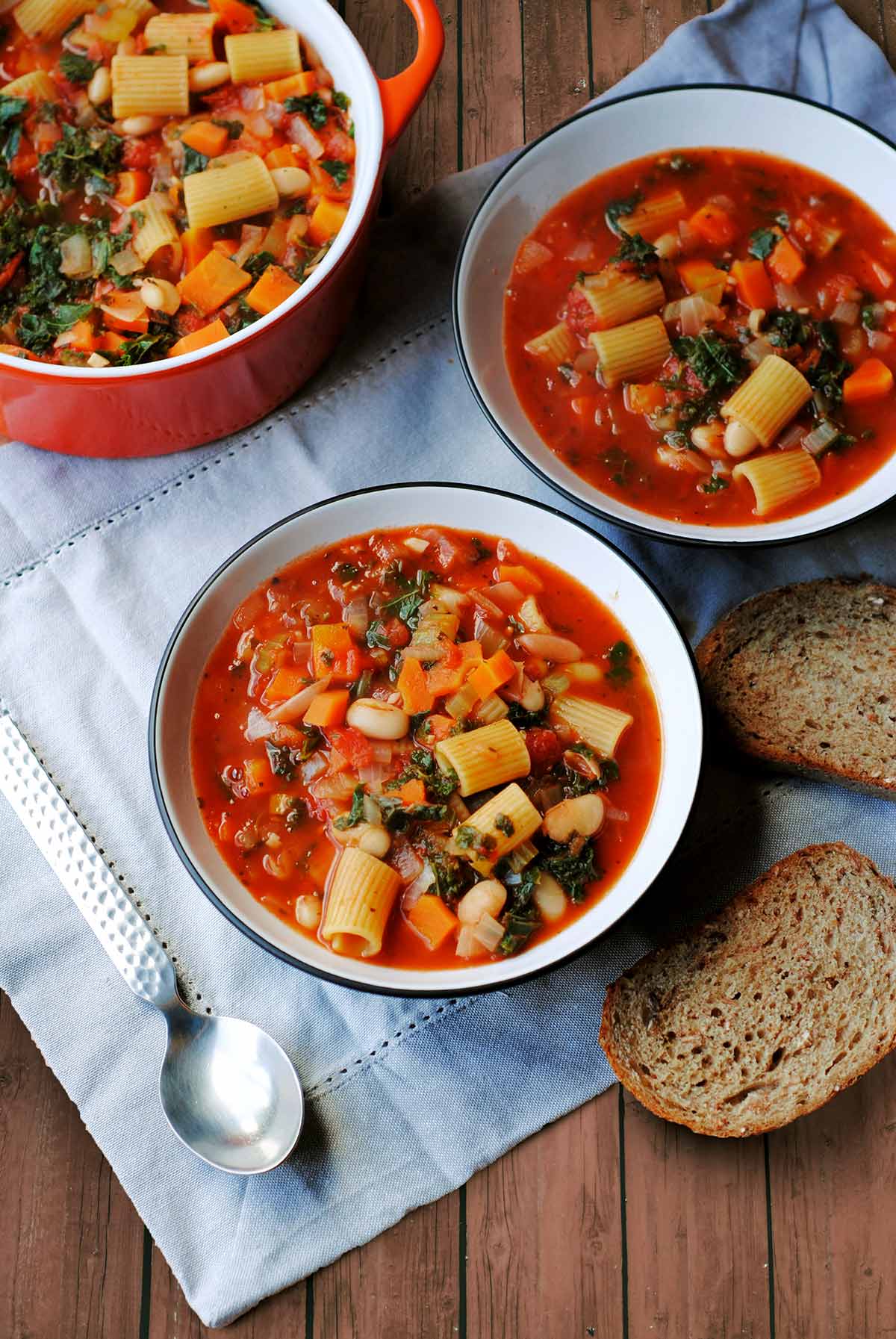 Two bowls of vegetable and pasta soup on a blue cloth next to bread and a spoon.