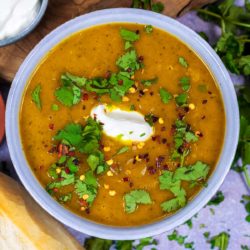 A bowl of carrot and coriander soup topped with cream, coriander leaves and chilli flakes.