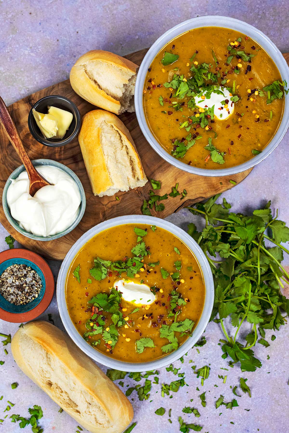 Two bowls of soup surrounded by bread and chopped herbs.