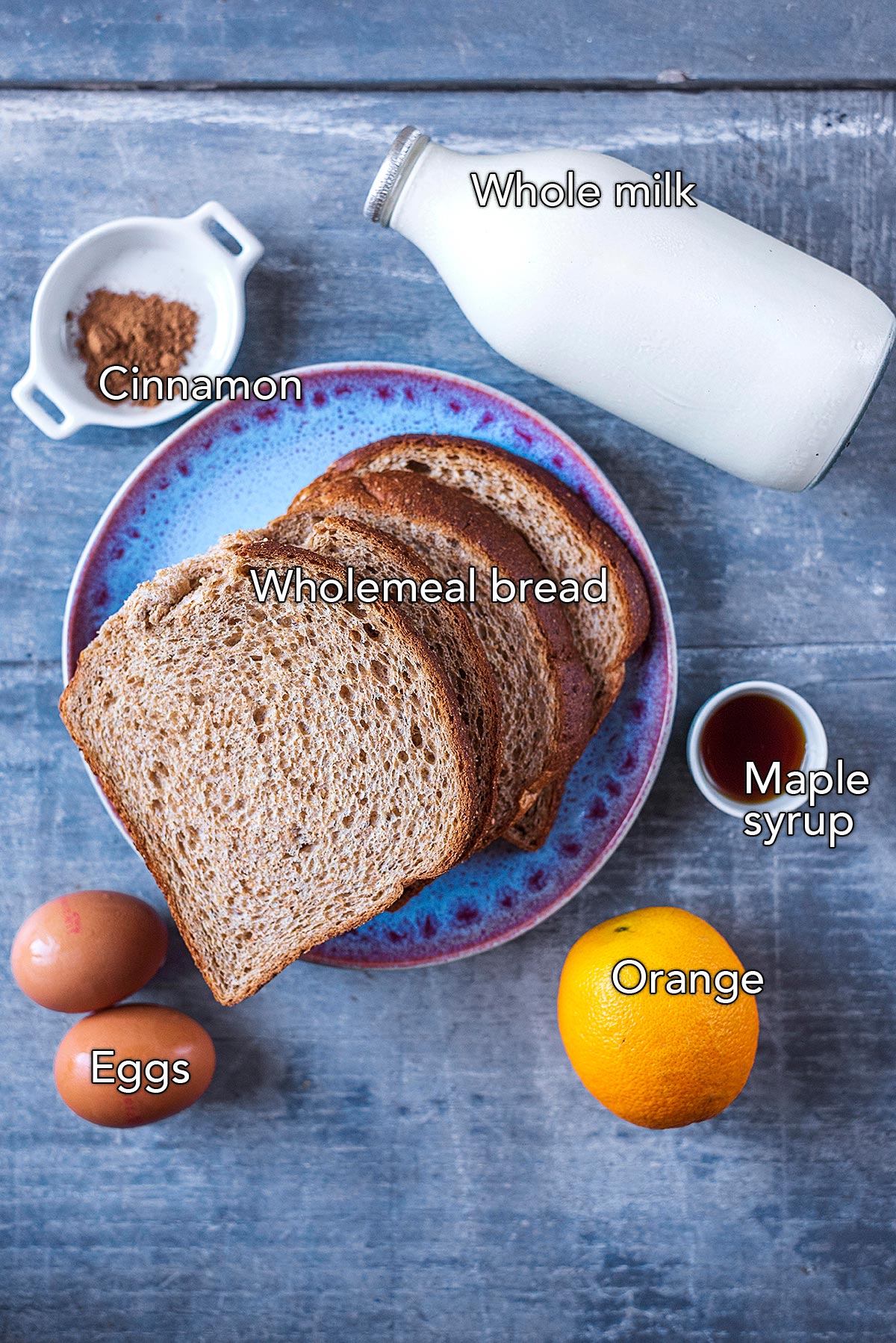Four slices of bread on a plate surrounded by a bottle of milk, two eggs, an orange, some maple syrup and some spices