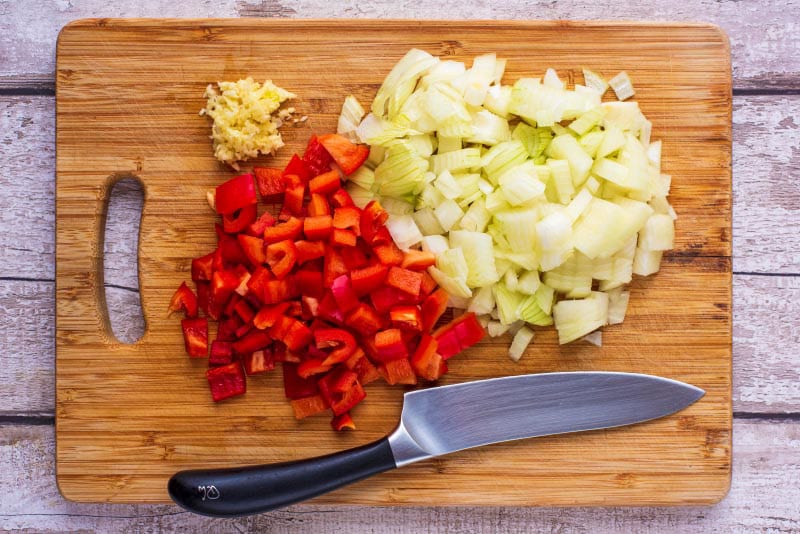A chopping board and chef's knife with chopped onions, red pepper and garlic.