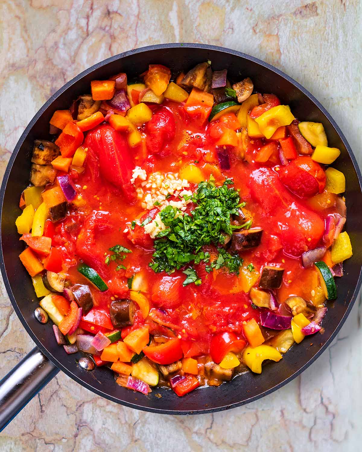 Chopped vegetables, plum tomatoes, chopped herbs and crushed garlic all cooking in a frying pan.