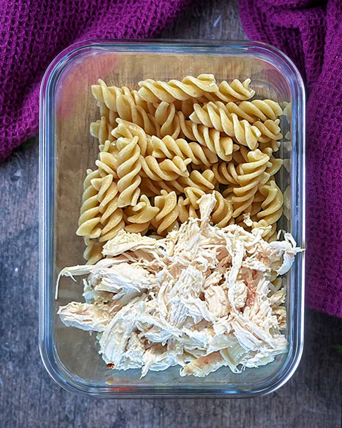 A glass container containing cooked fusilli pasta and cooked shredded chicken.