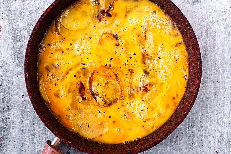 A frying pan with onions, potatoes and whisked egg cooking in it.