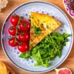A slice of Spanish omelette on a plate with vine tomatoes and green salad leaves