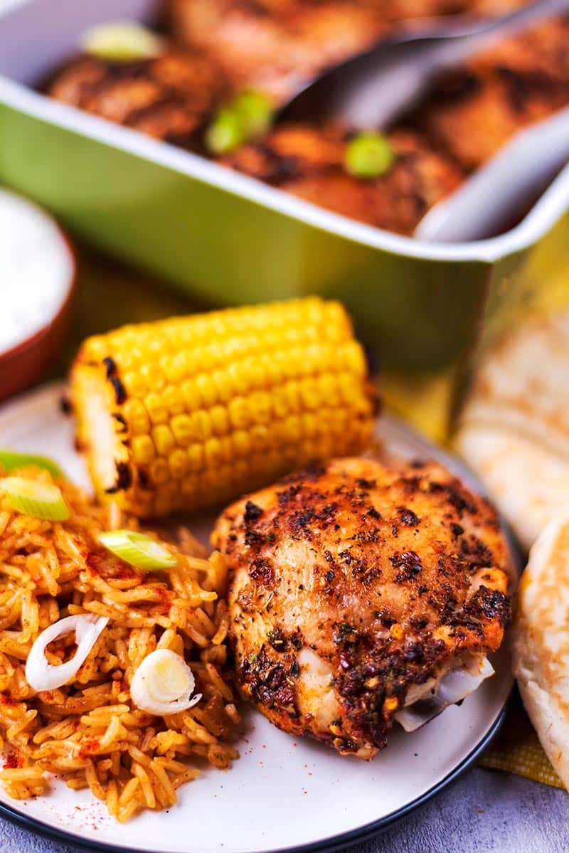 A chicken thigh, corn cob and rice on a plate in front of a baking dish full of chicken.