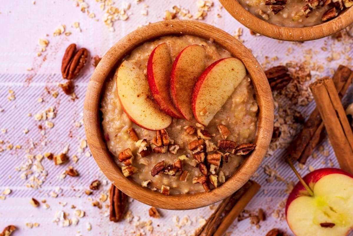 Apple Pie Oatmeal in a brown bowl with apple slices on top. Cinnamon sticks and an apple are next to it.