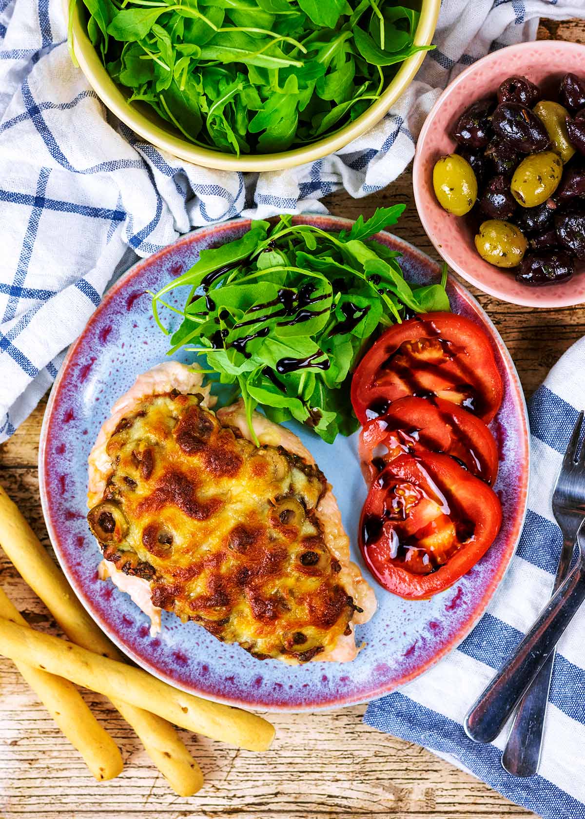 A cheese covered chicken breast on a plate with slices of tomato and some salad.