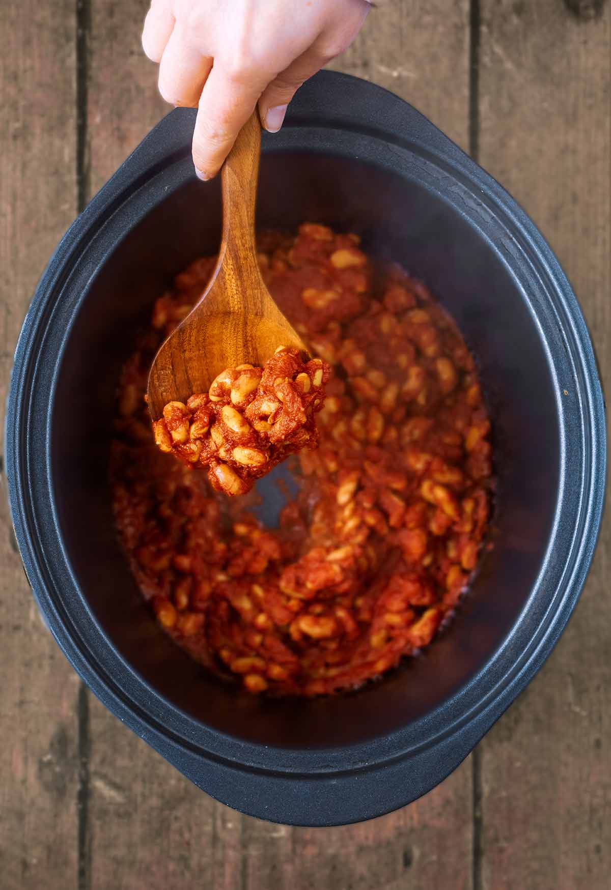 A hand holding a wooden spoon full of baked beans above a slow cooker pot.