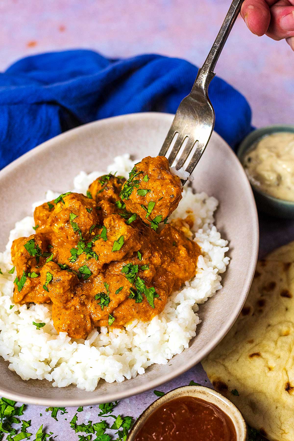 A fork picking up a chunk of chicken in a curry sauce from a bowl of curry and rice.