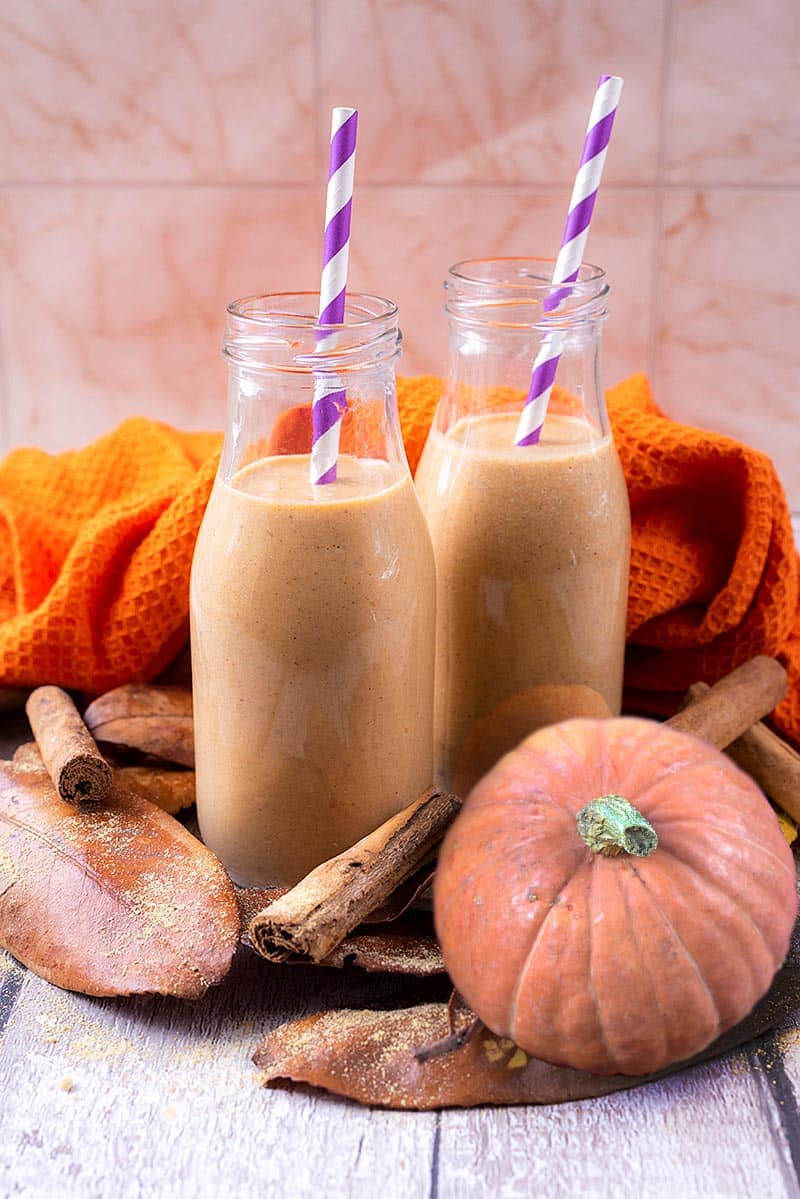 Two small milk bottles containing pumpkin smoothie with striped straws. A small pumpkin sits next to them.