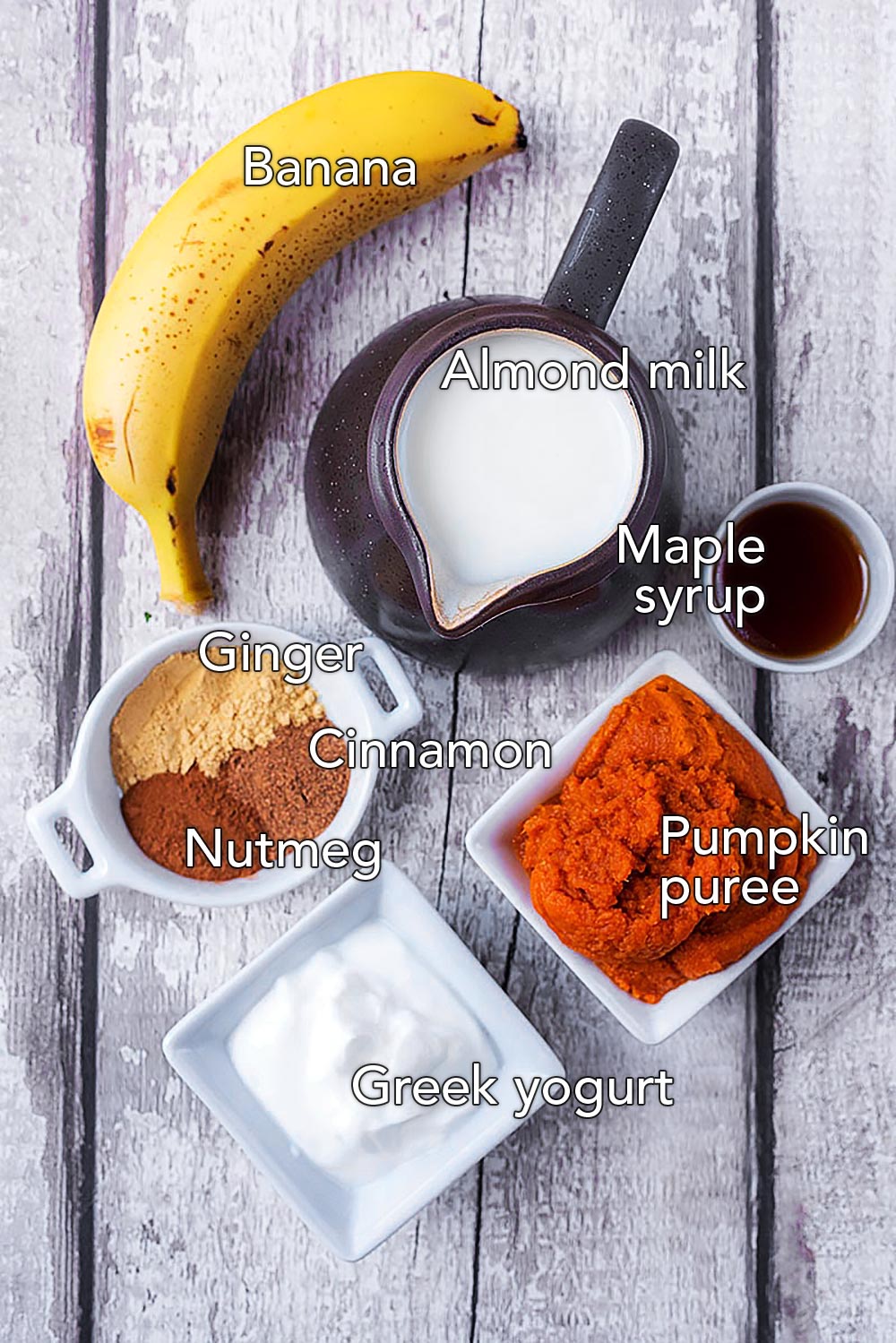 A banana, a jug of milk, some pumpkin puree, spices, maple syrup and yogurt all on a wooden surface.