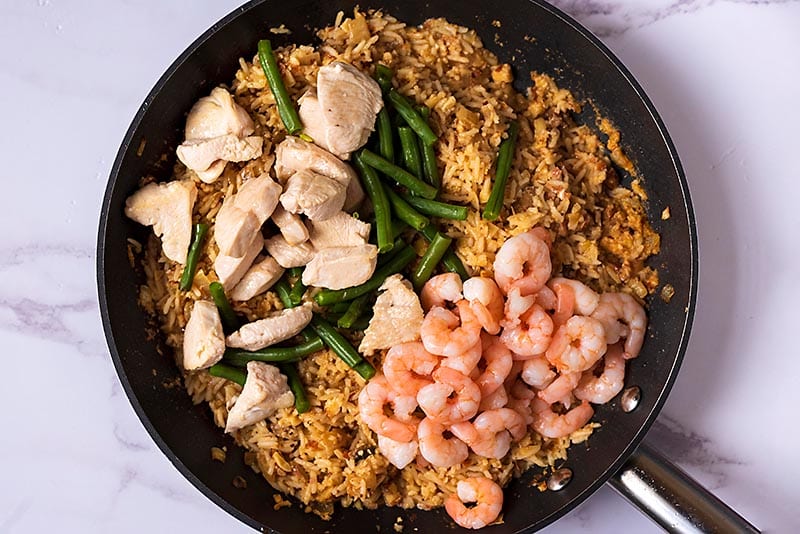 Fried rice, cooked chicken chunks, cooked shrimp and green beans cooking in a frying pan.
