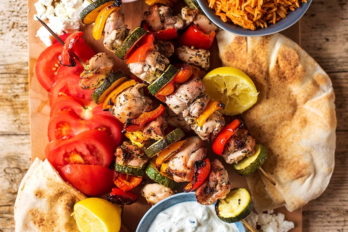 Vegetable and chicken skewers next to a bowl of rice, sliced tomatoes and some flatbreads.