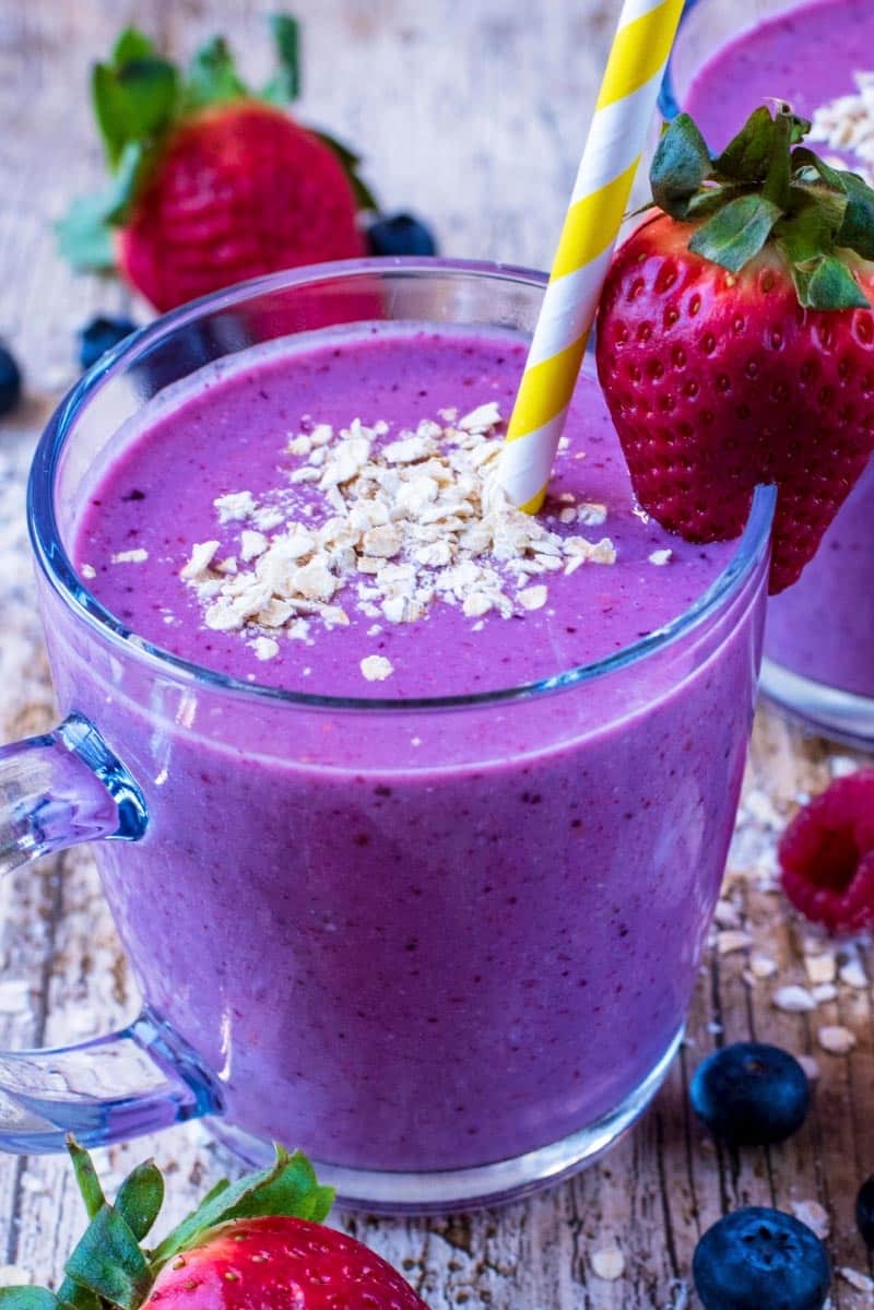 A glass mug containing a berry smoothie with oats floating on top and a strawberry on the rim of the glass