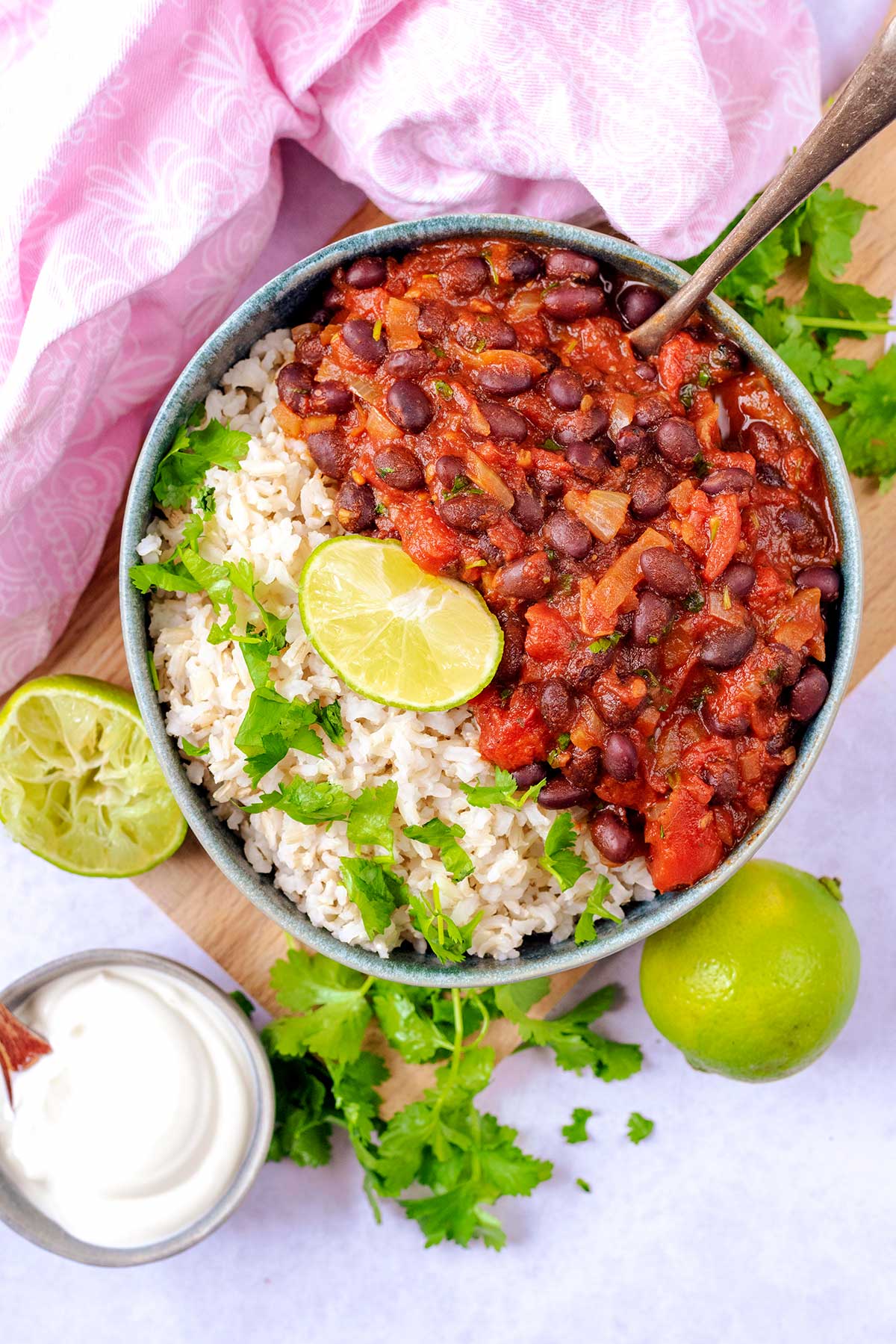 A bowl of bean stew and rice next to a small pot of cream, a lime and some coriander leaves.