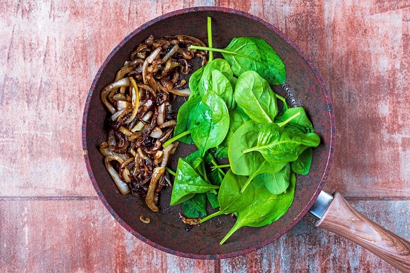 A frying pan containing sliced onions, chopped mushrooms and spinach.