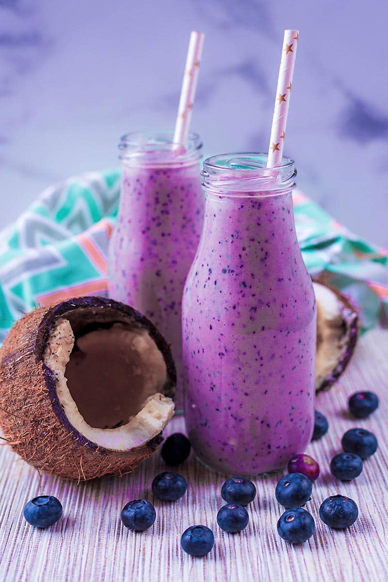 Two bottles containing a purple smoothie. Half a coconut and blueberries are next to the bottles.