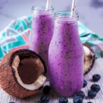 Two small glass bottles full of blueberry and coconut smoothie next to a broken open coconut