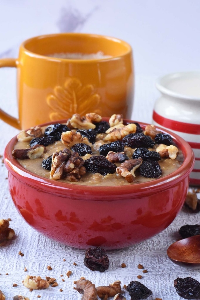 A bowl of oatmeal topped with walnuts and raisins with a cup of coffee in the background
