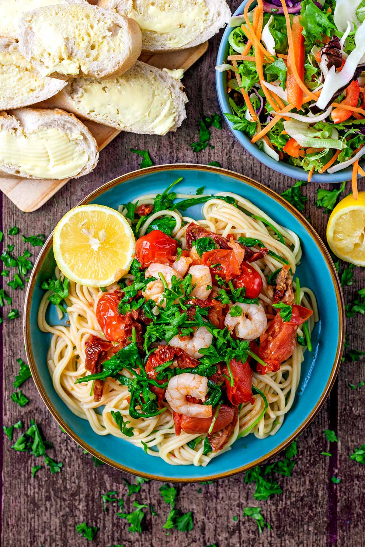 A plate of tomato and prawn spaghetti next to a board of bead and a bowl of salad.