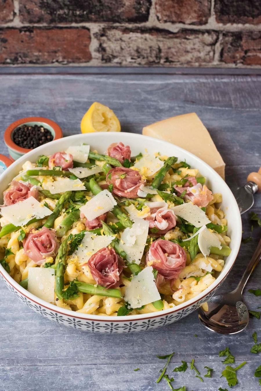 A bowl of asparagus and ham pasta next to a spoon, block of cheese, lemon half and pot of peppercorns.