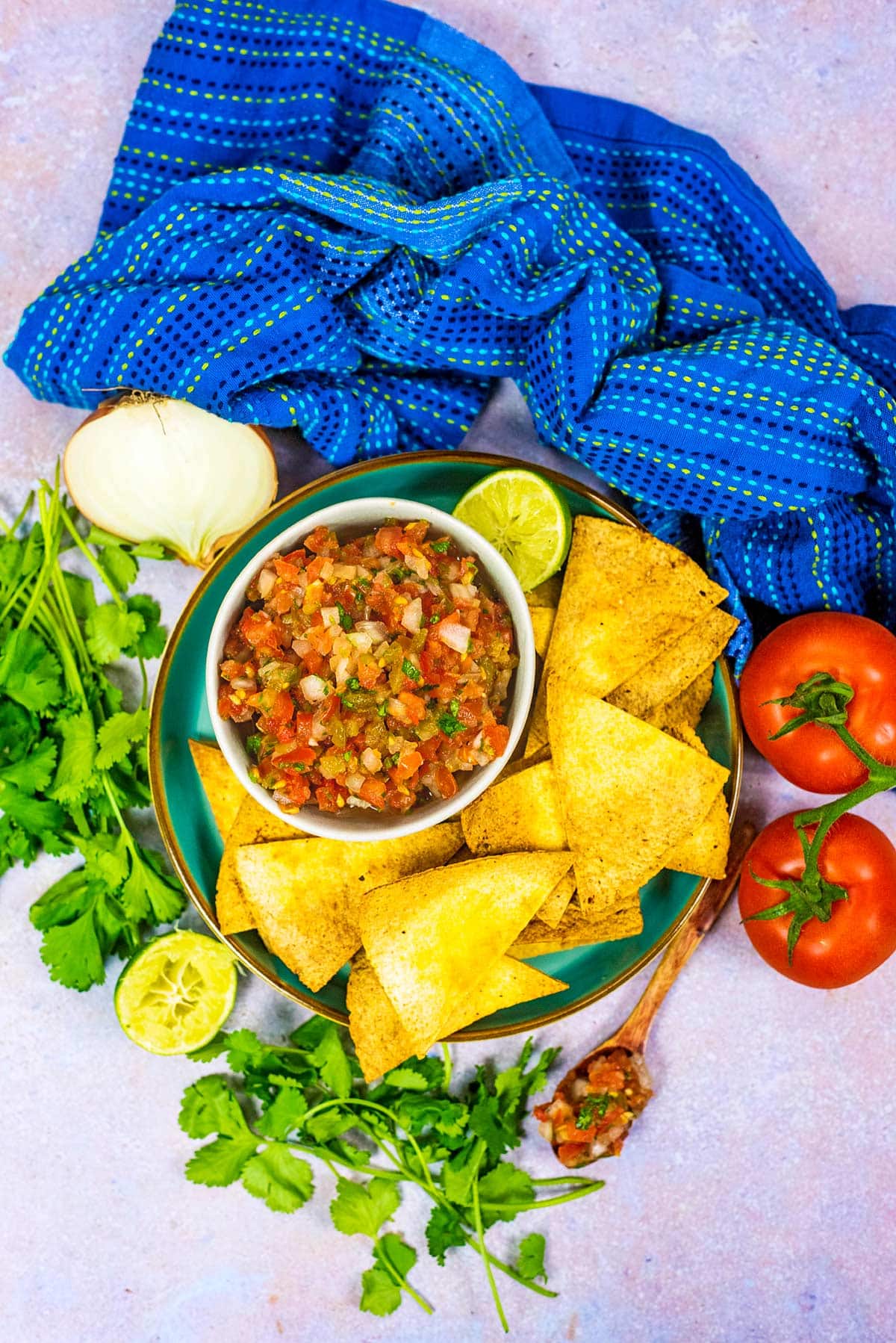 A bowl of salsa surrounded by tortilla chips, fresh coriander, tomatoes and lime halves.