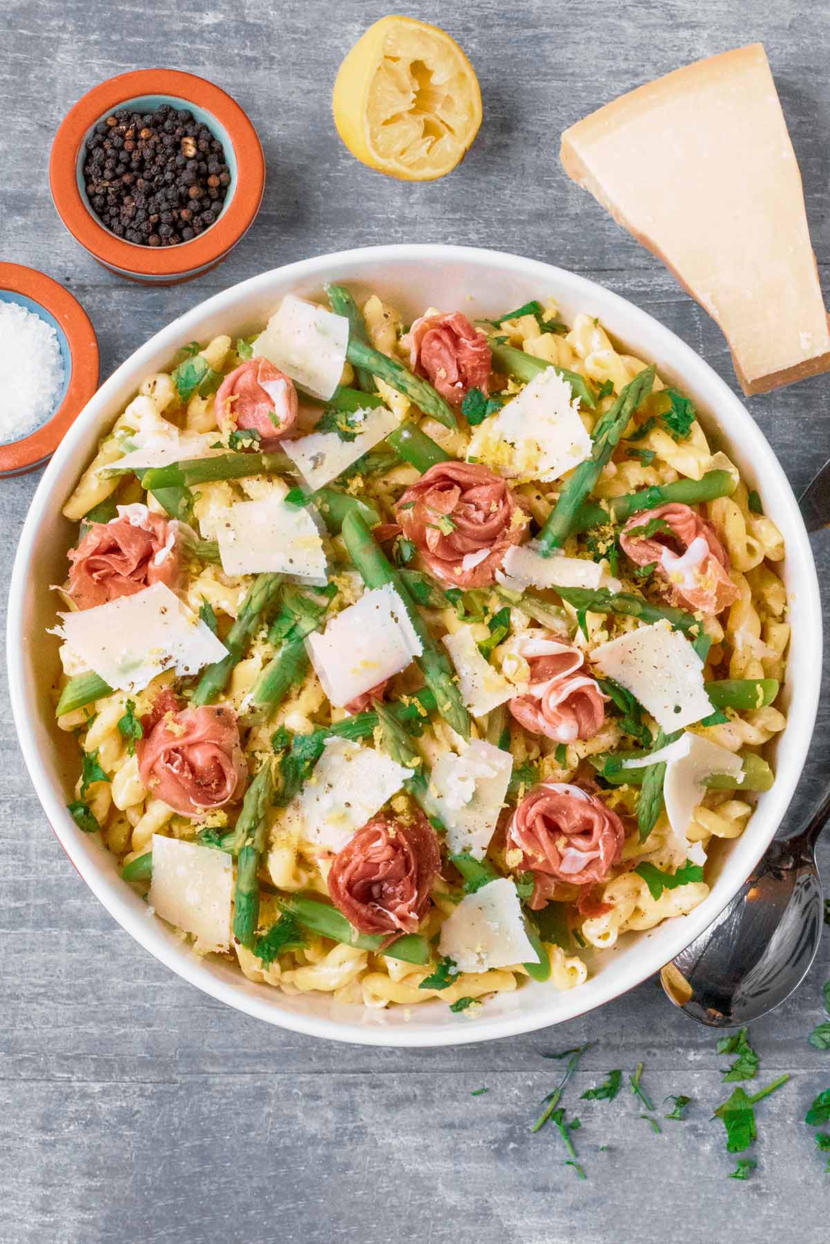 A bowl of asparagus and ham pasta next to a spoon, block of cheese, lemon half and pot of peppercorns.