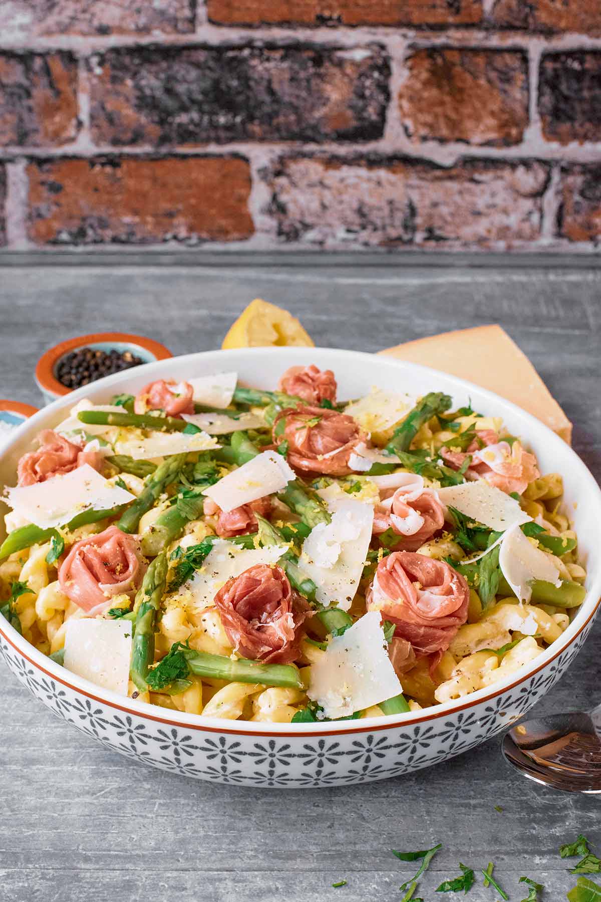 A bowl of ham and asparagus pasta in front of a brick wall.
