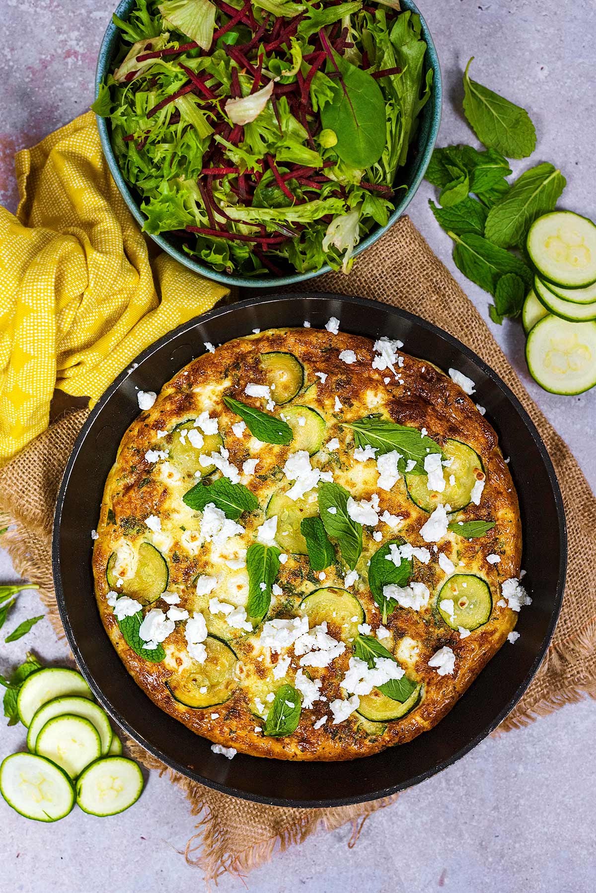 A frittata in a pan with a yellow towel wrapped around the handle. A bowl of salad is next to it.