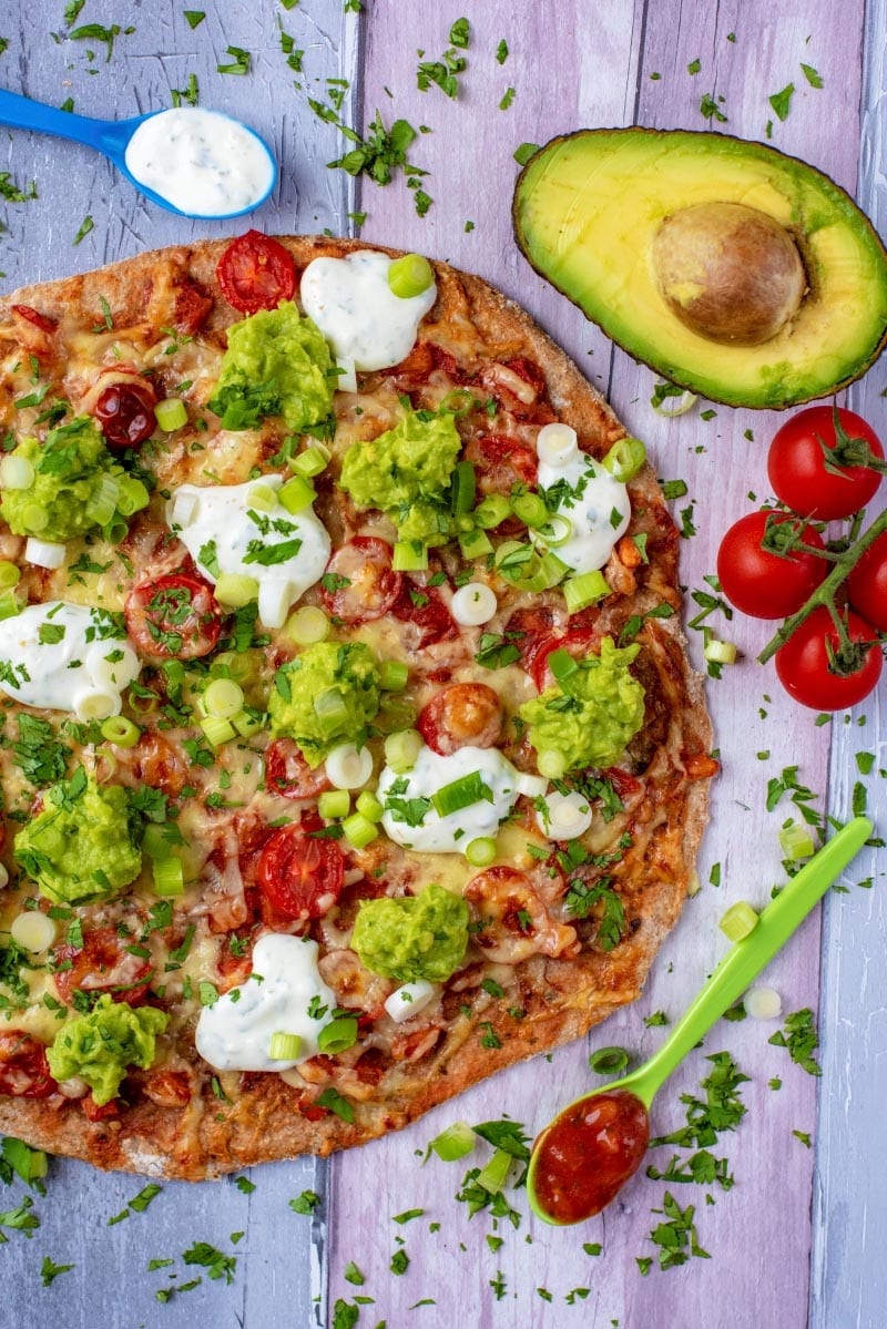 A pizza surrounded by avocado, tomatoes and spoons.