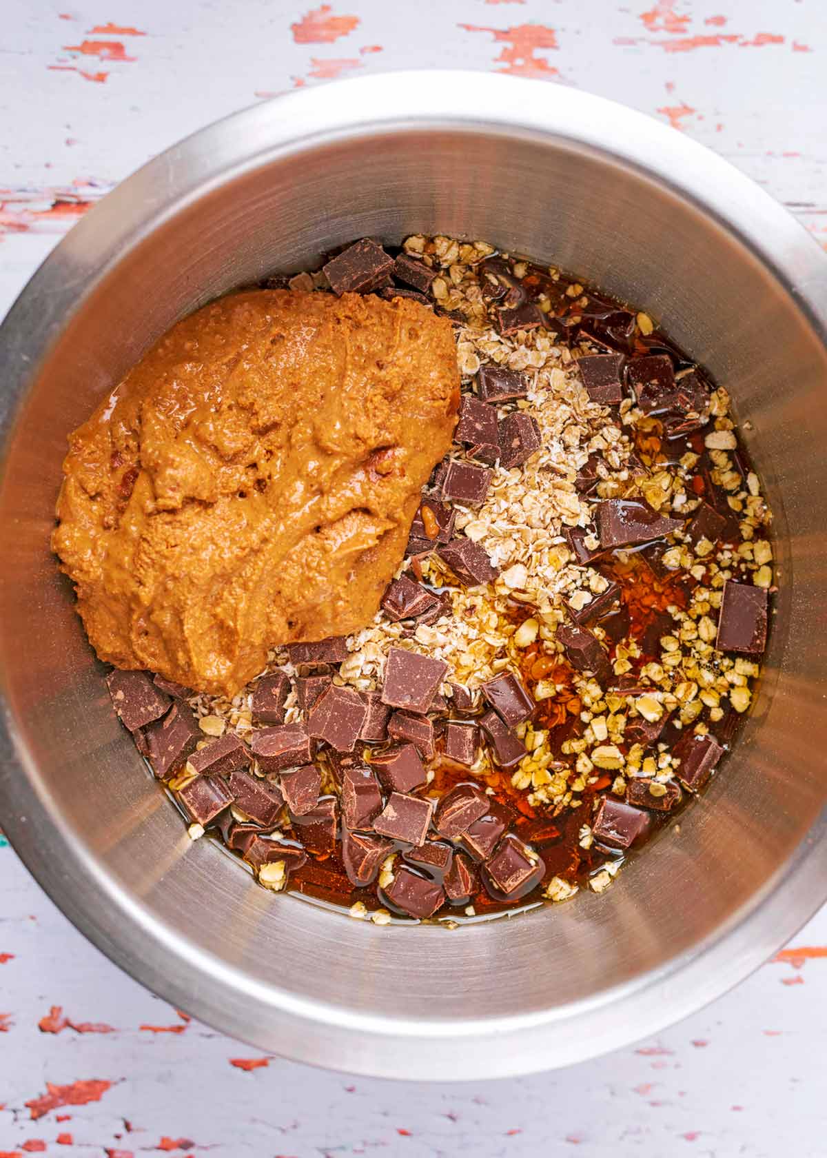 A mixing bowl containing oats, chocolate chunks, maple syrup and melted nut butter.