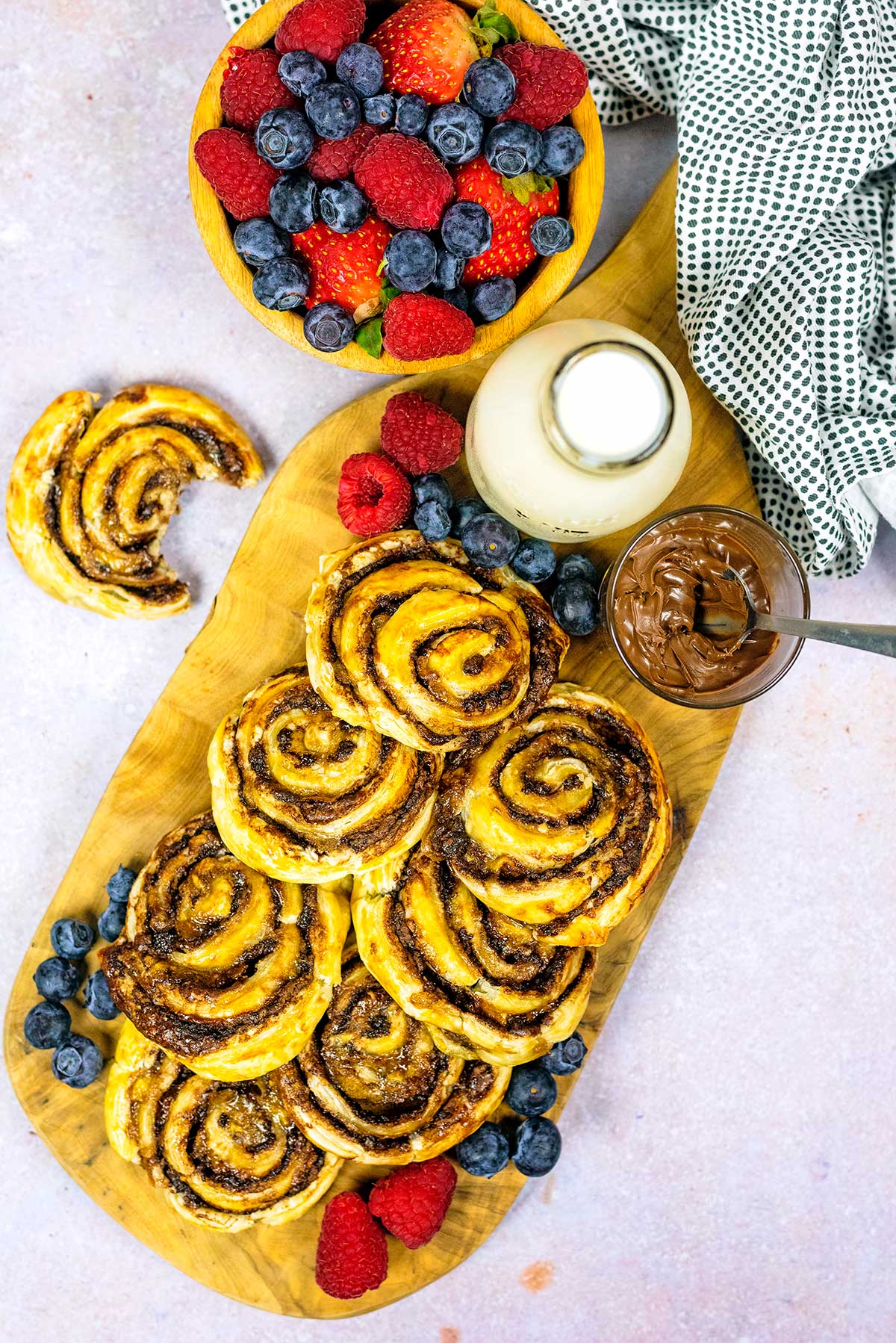 A wooden board covered in pinwheels with a bottle of milk and a bowl of berries.