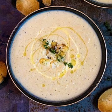 Creamy Roasted Cauliflower Soup in a bowl drizzled with cream and oil, topped with herbs.