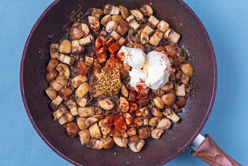 Cooked shopped mushrooms and shallots in a frying pan with dollops of cream and mustard. Paprika is sprinkled over everything.