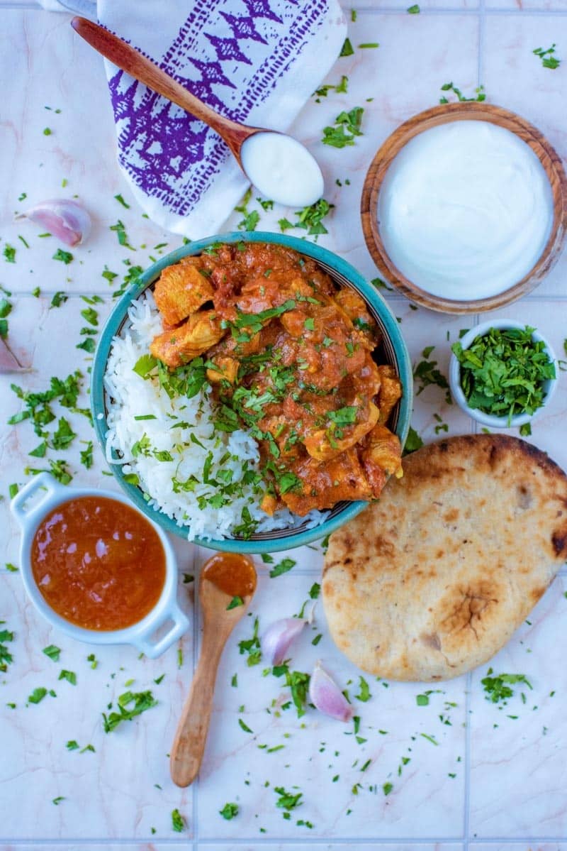 A bowl of curry and rice with a naan bread, sauces and herbs.