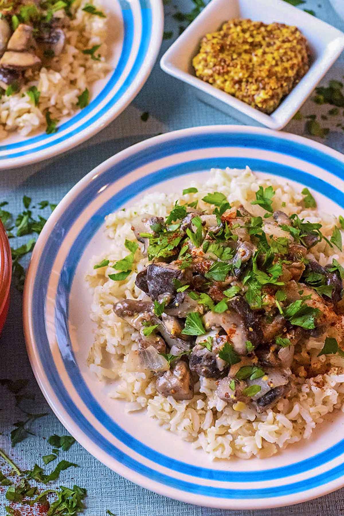 Mushroom Stroganoff served on two blue and white plates surrounded dishes of cream, mustard and rice.