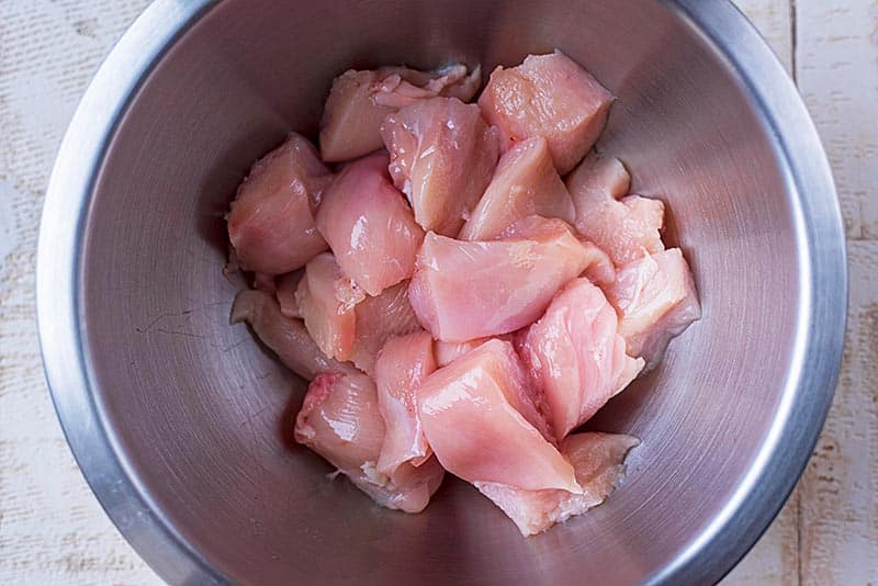 A large metal bowl containing chunks of raw chicken breast.