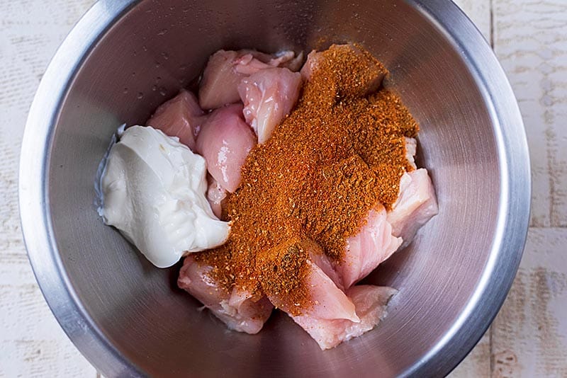 A metal bowl containing raw chicken breast chunks, some yoghurt and a spice blend.